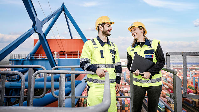 Zwei HHLA-Mitarbeitende in Sicherheitskleidung stehen auf einer Containerbrücke und unterhalten sich vor dem Hintergrund eines Containerterminals.
