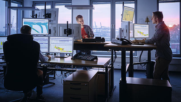 Mitarbeitende des Hamburg Vessel Coordination Center (HVCC) koordinieren Schiffsanläufe an Computerarbeitsplätzen mit Blick auf den Hafen.