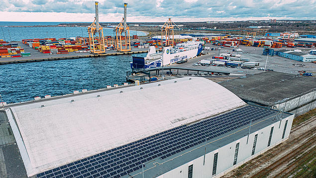 Luftaufnahme des Containerterminals HHLA TK Estonia im Hafen von Muuga mit großflächiger Solaranlage auf einem Terminalgebäude und Containern am Kai.
