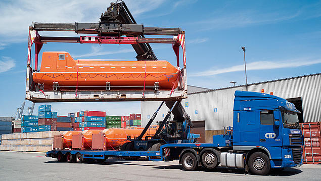 Schwergutlogistik auf der Straße: Ein blauer Lkw der HHLA-Tochter CTD wird mithilfe eines Reachstackers mit einem großen orangefarbenen Rettungsboot beladen. Die Szene zeigt die präzise Handhabung von Projektladung auf einem Terminalgelände.