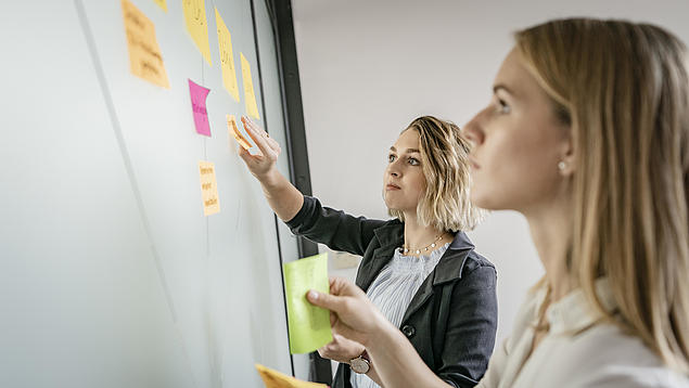 Zwei HHLA-Mitarbeiterinnen analysieren Markt- und Finanzthemen an einem Whiteboard mit Haftnotizen in einem Besprechungsraum.